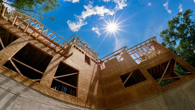 This image contains the wooden frame and red bricks structure for residential construction purpose while the sun is shining upon it in a beautiful blue sky, highlighting the structure.