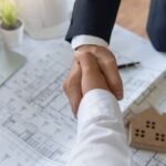 Two professionals shaking hands over architectural blueprints with a hard hat, model house, and laptop on the table, representing a construction agreement.