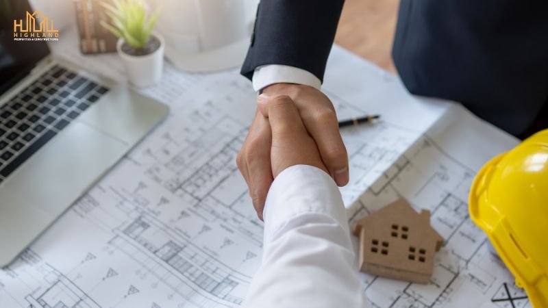 Two professionals shaking hands over architectural blueprints with a hard hat, model house, and laptop on the table, representing a construction agreement.