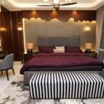 A luxurious bedroom interior design featuring a plush bed with a deep purple comforter, a striped bench at the foot of the bed, a stylish wooden desk, and elegant lighting. The room has a contemporary aesthetic with textured walls, floor-to-ceiling curtains, and a warm ambiance.