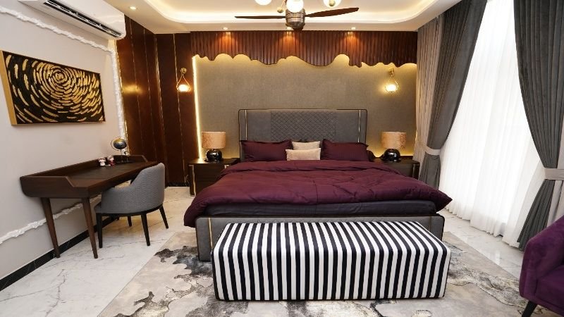 A luxurious bedroom interior design featuring a plush bed with a deep purple comforter, a striped bench at the foot of the bed, a stylish wooden desk, and elegant lighting. The room has a contemporary aesthetic with textured walls, floor-to-ceiling curtains, and a warm ambiance.