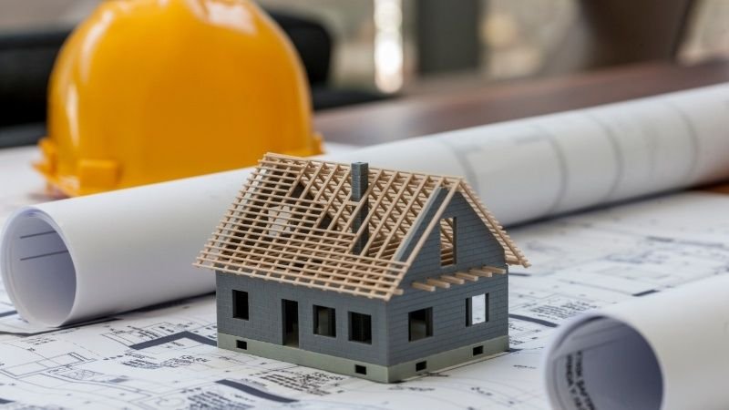 A miniature house model placed on architectural blueprints with a yellow safety helmet, representing House Construction Services in DHA Islamabad.

