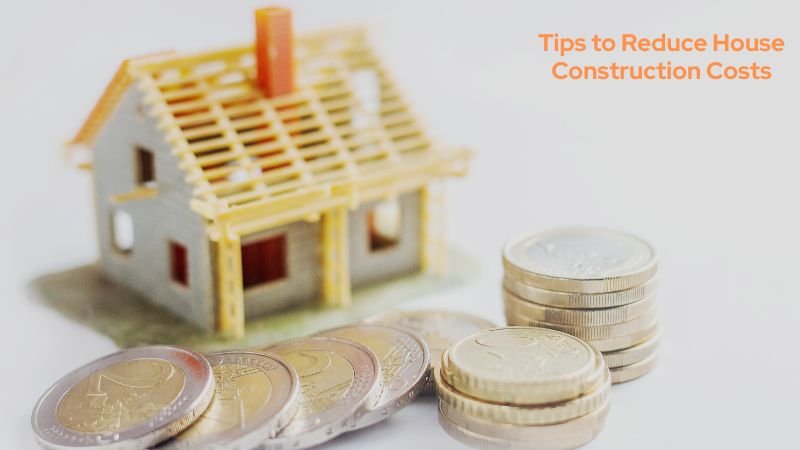 A miniature house model under construction with stacked coins in the foreground, symbolizing cost-saving strategies for home building.