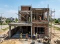 House Construction in DHA Lahore – A modern double-story house under construction with exposed brick walls, concrete framework, scaffolding, and construction workers actively working on-site in a residential DHA Lahore neighborhood.